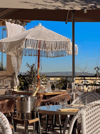 Terrasse de café avec parasol, tables en bois et vue sur un paysage ensoleillé.